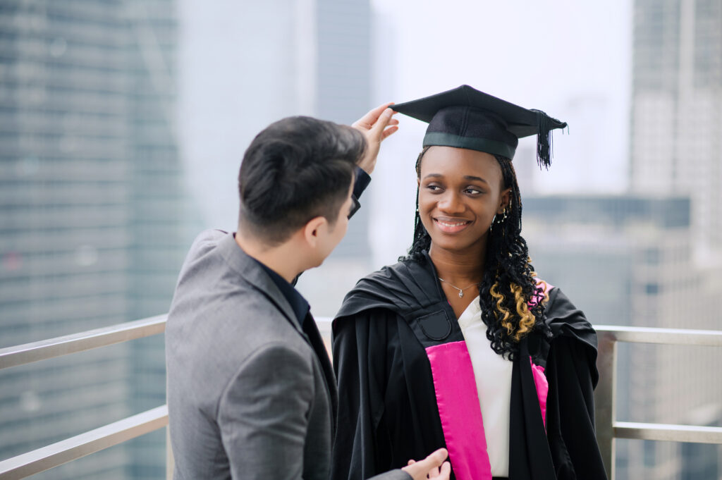 Boyfriend adjusting mortarboard for his girlfriend in college graduation gown outside college buildings getting ready for graduation ceremony.